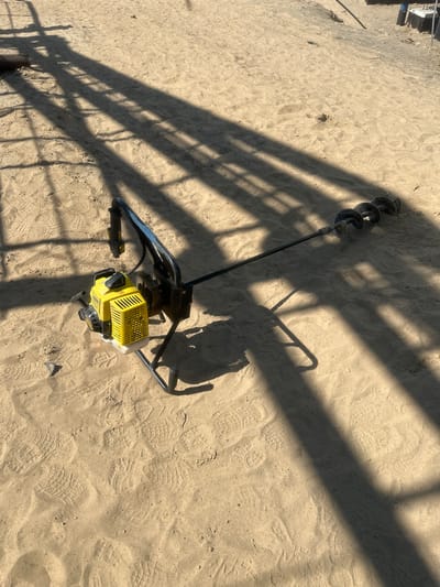 An auger is lying on the sandy ground in Чикшино, Russia. The auger is yellow and black and has a long, spiraled drill bit attached to it. The ground is covered in sand with numerous footprints visible. Long shadows are cast across the sand, indicating a sunny day. In the background, parts of a structure are visible, with its shadow also present.