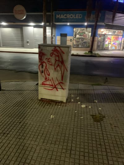 The image displays an urban street scene at night in Villa Bernardo de Monteagudo, Argentina. In the immediate foreground, a white utility box approximately 1.5 meters tall stands on a grey tiled sidewalk. The box is spray-painted with red graffiti depicting abstract, elongated figures. On the sidewalk around the box, several scattered, irregular white fragments, consistent with plaster or concrete debris, are visible, along with a dark, wet-looking stain on the tiles to the right.

Across the asphalt street, two commercial buildings are present. The building on the left features a partially visible circular sign with "SUPER" written in red, and two storefronts secured by closed grey roller shutters. The adjacent building to the right has a light blue facade with a white sign displaying "MACROLED". Its large glass display windows are brightly illuminated from within, showcasing various merchandise. A second illuminated storefront is visible immediately to the right of the MACROLED building. Overhead artificial lighting illuminates the street and storefronts. No individuals or vehicles are visible in the scene.
