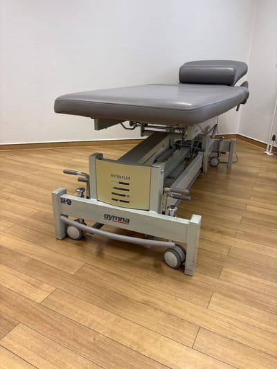 This is a medium shot, taken from a high angle, of a gray osteopathic treatment table on a light-brown wooden floor. The table is the main object in the frame and takes up most of the image. It appears to be a stationary piece of medical equipment, possibly found in a clinic or therapy room. The table is padded and appears to be adjustable, with various levers and mechanisms visible. The word "OSTEOFLEX" is printed in black text on a cream-colored panel on the side of the table. Below this, the word "gymna" is printed in red text on a white background. The floor is made of wooden planks laid horizontally, and the lighting suggests it is daytime indoors. There are no people visible in the image. The scene is calm and utilitarian, focusing on the medical equipment itself.