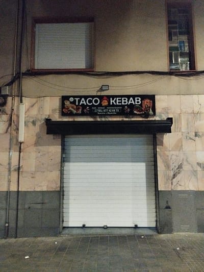 The image shows the facade of a closed "TACO KEBAB" restaurant in Rubí, Spain. A sign above the closed white roller shutter door displays the restaurant's name and a phone number. The building's facade is a combination of marble and grey paneling. Above the sign, there are two windows, one with a closed blind and the other with an irregular window pane design. Electrical wires and a white rectangular box are visible on the wall to the left of the sign. The pavement in front of the building is tiled.