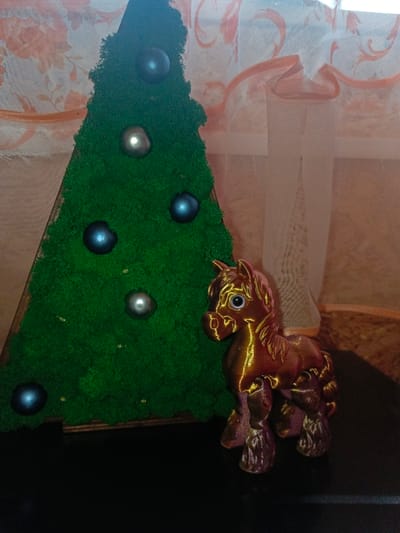 The scene is an indoor setting in Gorka, Russia, depicting two decorative objects arranged on a dark, reflective surface. To the left is a triangular, Christmas tree-shaped decoration, approximately 30-40 cm in height, covered in vibrant green, textured material resembling preserved moss. This decoration features five spherical ornaments: three dark blue and two silver-grey, distributed across its surface. To the right of the tree decoration stands a small figurine of a horse, approximately 15 cm tall. The horse figurine exhibits an iridescent or metallic finish, primarily reddish-brown with golden highlights, particularly on its raised features. It has prominent, round, light-colored eyes. The background consists of a window partially covered by two layers of curtains. The inner layer is a sheer, light-colored fabric, while the outer layer is a translucent, peach-toned curtain with a lighter, swirling floral pattern. No individuals are present, and no actions are occurring.