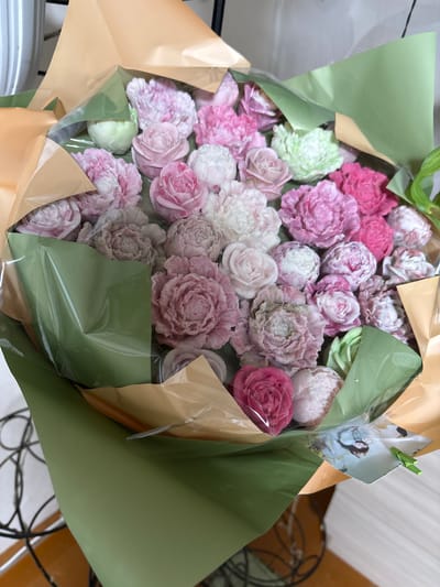 The image shows a close-up of a bouquet of soap flowers in various shades of pink, white, and green. The flowers are arranged in a bundle and wrapped in green and gold paper, with a clear plastic film covering the front. Some of the flowers resemble roses and peonies. In the lower right corner, a small printed card with an image of two people and a green clip is visible. The background is out of focus but appears to be an indoor setting with a white surface and some dark, wire-like structures. There are no people actively engaged in any actions within the frame. The specific location within Siberia, Russia, cannot be determined from the visual information provided.