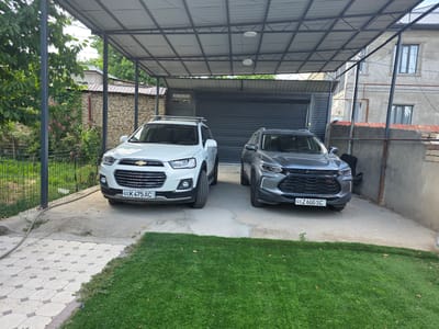 The image shows two cars parked under a carport in Yalangach, Uzbekistan. The carport has a metal roof and metal support poles. In front of the carport, there is a neatly manicured lawn. To the left of the carport, a brick wall is covered with ivy and some pink flowers. Behind the carport, a dark-colored garage door is visible. To the right of the carport, a two-story building with grey walls and windows can be seen. The license plate of the white car on the left reads "01 K 475 AC". The license plate of the grey car on the right reads "01 Z 600 SC". There are no people visible in the image.