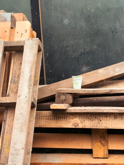 The image depicts a pile of wooden pallets stacked against a dark, textured surface, possibly a wall or a tarp. In the middle-right of the frame, a disposable paper coffee cup with a repeating "COFFEE" pattern in green text is balanced precariously on one of the wooden planks. The cup is white and appears to be empty. The pallets are weathered and show signs of rough use. The lighting suggests an outdoor or partially enclosed space. The scene is located in the city of Khelvachauri, Georgia.