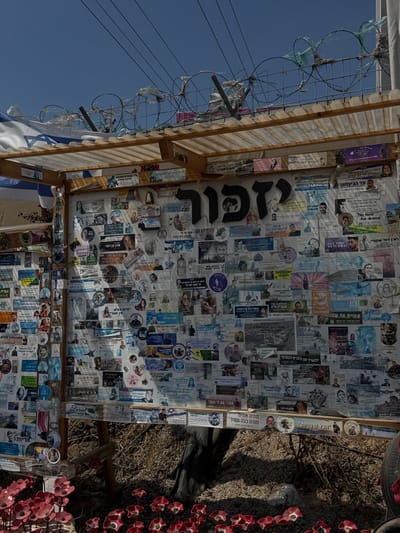 The image displays an outdoor memorial structure located in the Sdot Negev Regional Council, Israel. The primary element is a large, vertical panel within a wooden frame, topped by a translucent corrugated plastic roof panel. This panel is extensively covered with numerous printed papers, photographs, and stickers, predominantly featuring individual portraits and accompanying Hebrew text, indicative of memorial notices. A prominent, large black Hebrew word, "יזכור" (Yizkor), meaning "Remember," is affixed near the top center of the display.
Above the structure, coiled barbed wire is strung horizontally, with some plastic debris entangled within. A blue and white Israeli flag is partially visible on the upper left side, draped over the wooden frame. Utility lines are visible against the clear blue sky.
At the base of the display, the dry ground, covered in reddish-brown earth and sparse vegetation, extends into the foreground. Numerous artificial red flowers, resembling poppies, are arranged in rows across the ground. The scene is illuminated by direct sunlight, casting shadows. No individuals are present in the depicted area.