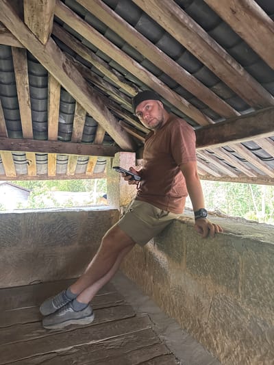 The image shows a man in Sà Phìn Commune, Vietnam, situated within an interior structure. He's wearing a brown t-shirt, khaki shorts, grey socks and shoes, and a black cap. He is holding a small black device in his left hand and is propping himself up against a rough stone or concrete wall with his right hand. He is wearing a large black watch on his left wrist.
To his left, wooden beams and a tiled roof can be seen. Through an opening or window, greenery is visible in the background. The ground appears to be made of wood planks or stone. The overall impression is that the man is exploring or inspecting an old structure.