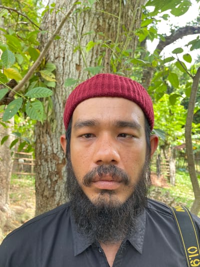 A man with a beard and a red beanie is standing in front of a large tree, with green foliage and sunlight filtering through the leaves in the background. A yellow strap with the text "Nikon" is visible across his chest. The man is looking directly at the camera. The scene appears to be outdoors, possibly in a park or natural setting. The location is identified as Seulimeum, Indonesia.