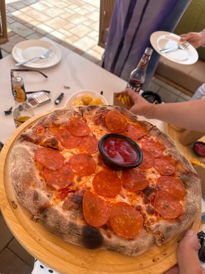 A person is holding a wooden pizza peel with a pepperoni pizza on it. The pizza has a thick crust and is topped with a generous amount of pepperoni slices. A small black bowl of ketchup is placed in the center of the pizza. In the background, a table is set with plates, forks, a bottle of soda, and a pack of cigarettes. Another person's hand is visible reaching for a plate. The scene appears to be outdoors, with a casual setting, possibly a restaurant or a picnic. The time of day seems to be daytime. The overall mood is relaxed and social, suggesting a meal being shared. A bottle of soda has some text on it, but it is not clearly legible. The location is identified as Ptghni, Armenia.