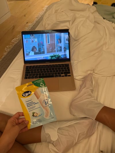 In Vila Nova de Cerveira, Portugal, an indoor scene depicts a person engaged in self-care and entertainment. A gold Apple MacBook Air laptop rests open on white, rumpled bedding, displaying an interior scene from a video featuring multiple individuals and window blinds. The person's lower legs and feet, with a tanned complexion, are visible. Both feet are encased in white, single-use foot masks. The person's left hand holds a yellow and light blue package of "Scholl PediMask Intensely Moisturises, Repairs, Softens," which indicates "1 Pair" and contains avocado oil. A light-colored wooden floor and a textured gray rug are visible in the background to the left.