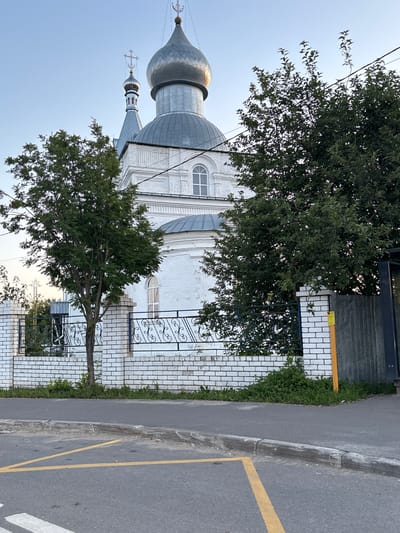 The image shows a white brick church with a silver dome and a smaller dome behind it, both topped with crosses. The church is partially obscured by two trees with green foliage. In front of the church is a white brick wall with a black wrought-iron fence. The area in front of the fence is a paved surface, likely a sidewalk or road, with yellow markings. To the right of the image, a grey metal fence is visible. The sky is clear and blue. The scene appears to be in a calm, outdoor setting. The location is identified as Bogorodskoe, Russia.