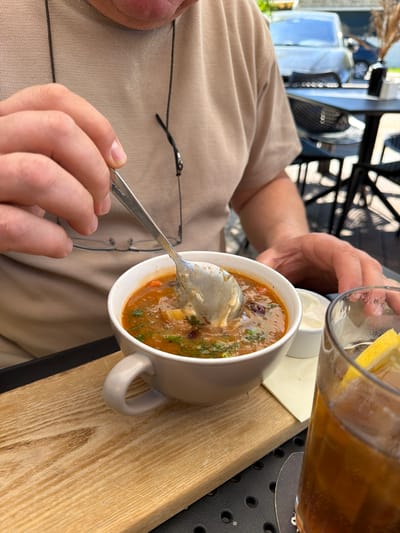 A person is seated at an outdoor table, holding a spoon in their right hand and a glass containing a beverage (possibly iced tea with lemon) in their left hand. The person is eating soup from a white mug. The soup contains herbs and other ingredients. A small white ramekin sits next to the mug. The table is a light-colored wood, set on a black wrought iron base. The scene takes place in Ragaciems, Latvia. Eyeglasses rest on the person's beige shirt.