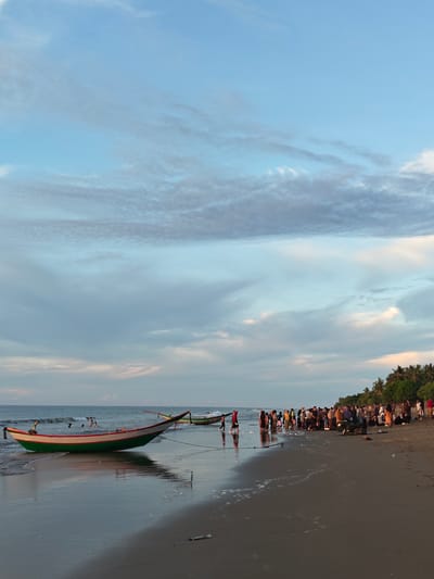 In the city of Uleekareung, Indonesia, a group of people are gathered on a sandy beach. The sky above is a mix of blue and grey clouds, indicating either dawn or dusk. The ocean is calm with gentle waves lapping the shore. In the foreground, a colorful boat with green and white stripes rests on the wet sand. Further down the beach, a large crowd of individuals stands near the water's edge, some appearing to be involved in activities related to the sea or boats. There are several other boats visible in the distance. The environment is characterized by the beach, the sea, and lush green trees lining the horizon.