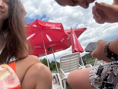 In Shipkovo, Bulgaria, four individuals are captured in a low-angle, close-up photograph, appearing to be at an outdoor seating area, possibly near a pool or resort. The scene is dominated by two large, red Coca-Cola branded umbrellas, providing shade.
In the foreground, on the left, a person's head and shoulder are visible. Their long, brown hair is partially obscuring their face, and they are wearing an orange and white patterned towel or sarong. To their right, another person's legs are visible, wearing patterned swim trunks with the letters "NKE" on a tag. Their right arm is extended, holding a smartphone.
Above and behind the foreground figures, the heads and faces of two other individuals are visible, looking down towards the camera. One has dark hair, and the other's face is partially obscured by their hand.
In the background, white plastic lounge chairs and a metal railing are visible. Beyond that, lush green trees and a cloudy sky form the backdrop. The presence of umbrellas and lounge chairs suggests a recreational setting, likely during daylight hours given the illumination and cloud cover. The photograph is taken from a perspective that emphasizes the subjects and their immediate surroundings.
