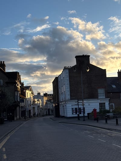 The image shows a street scene in Dacorum, United Kingdom, under a sky filled with clouds. The photograph is taken from a low angle, looking down the street. Buildings line one side of the street. The building closest to the camera is two stories tall and has a dark brick facade, with a light-colored lower section. A red post box is positioned on the sidewalk in front of it. The street is paved with brick and marked with yellow lines. The road surface is dark and appears to be wet. The sky is visible above the buildings, filled with cumulus clouds. A contrail from a jet aircraft is visible in the upper left.