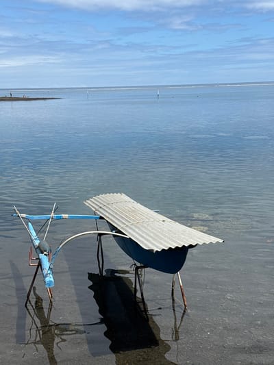 The scene depicts a calm, shallow coastal area in Māhina, France, under a clear sky with scattered white clouds. In the foreground, partially submerged in clear, blue-green water, are two improvised structures. The structure on the left consists of a blue, log-like beam supported by rusty metal poles forming a tripod, with a smaller blue cross-beam and a dark cylindrical object attached. The structure on the right features a dark blue, inverted trough or hull-like object covered by a corrugated galvanized metal sheet, secured by rope, and also supported by rusty metal poles. Both structures cast dark, elongated shadows onto the visible sandy seabed. In the midground, the calm water extends, revealing two slender white poles protruding vertically. Further in the background, a low-lying landmass or sandbar is visible on the left, with three indistinct human figures standing on it. Beyond this, a faint white line indicates a distant reef or wave break, separating the calm water from the open sea under a hazy horizon.