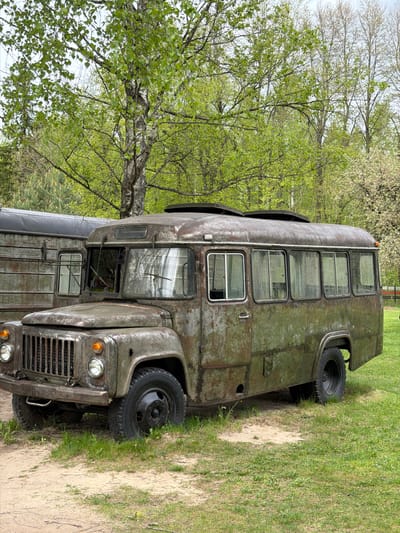 An antiquated, military-style vehicle sits partially on a sandy/dirt path and partially on a grassy area. The vehicle appears to be a bus or large truck with a boxy structure and heavily rusted or weathered exterior. The front features a grill, headlights, and a windshield with a wiper. The side of the vehicle is punctuated by several rectangular windows. The tires are black and appear to be standard truck tires. In the background, there are trees with green leaves, suggesting the scene is set during a temperate season. The environment seems rural or suburban, rather than a dense urban setting. The location is Salaspils, Latvia.