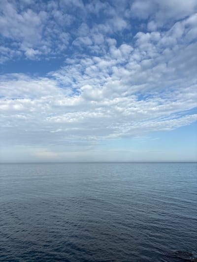 The image depicts a stark horizontal division between sky and sea, possibly in Bondarenkovo, Ukraine. The left side of the image is dominated by a vibrant blue sky filled with white, puffy clouds arranged in elongated formations. Moving towards the center, the sky fades to a lighter, hazy blue, nearing the horizon. On the right side, the sea extends to the edge of the frame, its surface a uniform, slightly rippled texture. The water appears greyish-blue, possibly reflecting the overcast sky. The horizon line, separating the sky and the sea, is a distinct, straight boundary. No people, objects, or specific actions are visible.