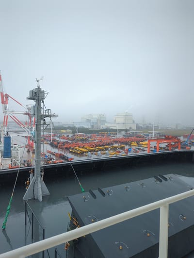 This image depicts a view of the port of Tōkai, Japan, on a cloudy, rainy day. In the foreground, a dark, metallic structure with white railings is visible, likely part of a ship or dock. The water is dark and reflects the overcast sky, with visible raindrops creating ripples on its surface.

In the midground, a large dock area is filled with a vast quantity of heavy machinery, predominantly yellow and orange excavators and dump trucks, neatly arranged. A large, dark gray barge or ship is docked alongside the pier. Further back, industrial buildings, including warehouses and a power plant with a smoking chimney, are visible. Cranes, both large red and white structures and smaller blue ones, are scattered throughout the port, indicating ongoing operations. The sky is heavily overcast and foggy, obscuring the distant horizon. A few figures, presumably workers, are discernible near the docked barge. The overall impression is a busy, industrial port environment operating under inclement weather conditions.