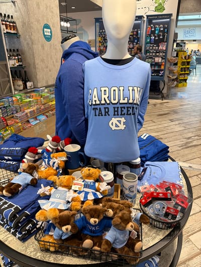 The image shows a retail display of merchandise, likely in a gift shop or convenience store, with a focus on University of North Carolina (UNC) apparel and souvenirs. The primary subjects are several UNC-themed t-shirts and stuffed teddy bears wearing UNC clothing. There are also miscellaneous items like bottle openers, magnets, and mugs.

The scene is an indoor setting with a wooden floor. The merchandise is arranged on a tiered display table with black metal baskets and shelves. In the background, shelves stocked with various products, including snacks and toiletries, are visible, along with signs for "Pamper Yourself" and "Baked From Scratch." A small section of a cooler or refrigerated display with bottles can be seen on the lower right.

The overall impression is a clean, well-lit retail environment. No specific time of day or weather conditions can be determined. The UNC branding suggests a location near the university or a store catering to fans. The items are new and packaged, indicating a commercial sale. Visible text includes "CAROLINA," "TAR HEEL," "DUKE," "LOCAL," "BAKED," "From Scratch," "MAGNET," and "BOTTLE OPENER." There are also price tags with "$8.99."