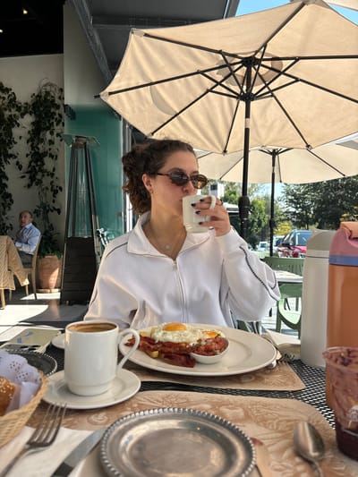 A person in sunglasses drinks from a mug at an outdoor table under an umbrella. A plate with eggs, bacon, and salsa sits nearby, along with a cup of coffee, utensils, and a bread basket. A man is seated further back. The background includes greenery, a glass panel, and a partial view of other tables and buildings. The overall setting suggests a casual outdoor dining experience.