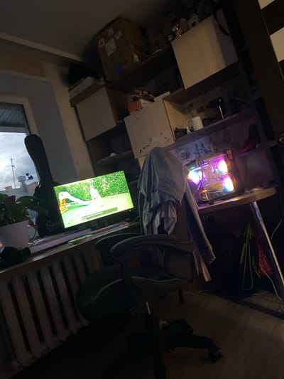 The image depicts a cluttered room, likely a home office or gaming setup, with a desk area prominently featuring a computer and a monitor. The computer tower is illuminated with colorful RGB lighting, suggesting it is powered on and possibly in use. On the desk, a monitor displays a video of a goose on a golf course. Various objects are scattered around, including boxes, what appears to be a gym bike, and some clothing or fabric draped over a chair. The lighting in the room is dim, with the computer's lights and the monitor being the main sources of illumination, indicating it is likely evening or night. The overall impression is one of a lived-in space filled with electronics and personal belongings. There is no visible text in the image.