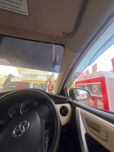 The image captures a scene from within a Toyota vehicle, identified by the steering wheel emblem, parked at a Shell fuel station in Abbottabad, Pakistan. The perspective is from the driver's seat, looking forward through the windshield and towards the passenger side during daylight hours.
Inside the vehicle, the black steering wheel, a dashboard displaying a speedometer marked to 220 km/h, and an ignition with keys are visible. A beige-colored air vent is present on the passenger side dashboard, and an overhead sun visor contains a printed warning regarding child seat safety. A dark weather deflector is affixed to the passenger side window frame.
Outside, under a Shell-branded canopy, a fuel station attendant, wearing a red uniform shirt, a yellow reflective vest with Shell logos, and a red cap, is actively dispensing fuel into a silver sedan positioned to the left of the viewer's vehicle. An additional individual, dressed in a dark cap and shirt, stands near the silver sedan. To the right, outside the passenger window, a Shell display cabinet branded "Shell Helix Ultra" showcases "Shell Rimula" oil products, with multiple QR code posters on its side. A masked person is discernable in the reflection of this display cabinet. A multi-story building with yellow trim is visible in the background beyond the immediate fuel station infrastructure.