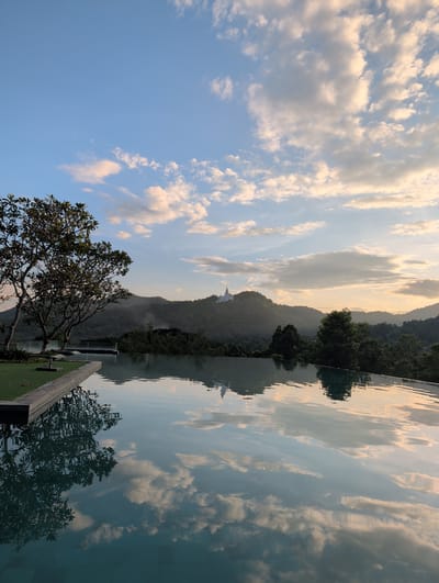 The image depicts a serene, panoramic view from an elevated perspective, likely a balcony or infinity pool, overlooking a lush, mountainous landscape in Ban Pong Nuea, Thailand. The scene is set during the late afternoon or early evening, as evidenced by the warm, golden light of the setting sun casting a soft glow on the scattered, fluffy clouds. The sky is a gradient of pale blue and soft orange, creating a tranquil atmosphere.

In the foreground, to the left, a large, leafy tree with a gnarled trunk stands prominently, its branches reaching out towards the sky. To the right, a reflective body of water, possibly an infinity pool, mirrors the sky, clouds, and the distant mountains. The water's surface is gently rippled, creating a shimmering effect on the reflections.

Beyond the immediate foreground, rolling hills covered in dense green vegetation dominate the mid-ground. A faint mist seems to shroud the lower slopes of the mountains, adding to the ethereal quality of the scene. On one of the higher peaks, a white structure, likely a temple or stupa, is visible, subtly illuminated by the fading sunlight.

The overall mood is one of peace and natural beauty. The image captures a moment of quiet contemplation, with the natural elements of sky, water, and mountains harmonizing to create a picturesque setting. There are no people or specific events depicted; the focus is purely on the tranquil landscape and the interplay of light and reflection.