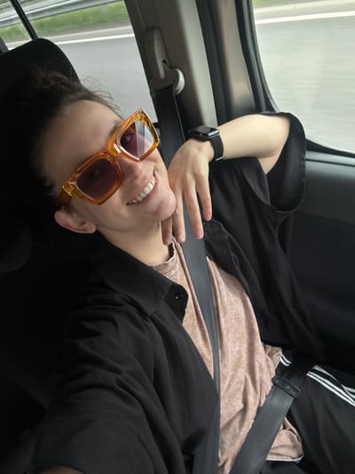 A person wearing orange sunglasses and a black watch is shown seated in a car, likely in transit. The person is wearing a black jacket and a light brown shirt underneath. A seatbelt is fastened across their body. The background shows a blurred view through the car window, suggesting motion. The location, Shali, Russia is not verifiable from this image.