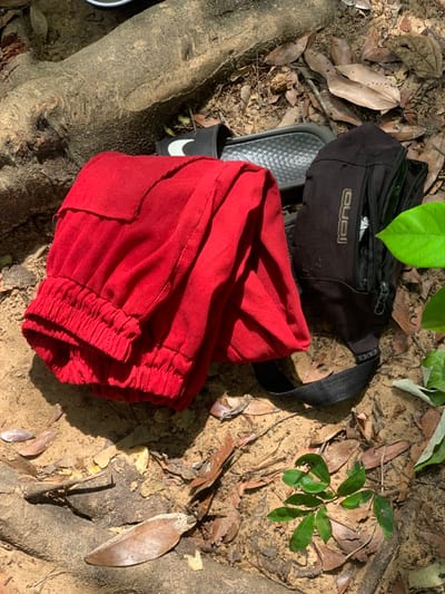 A pair of red corduroy pants are lying on the ground in Egbeda, Nigeria, near a black fanny pack and two pairs of flip-flops. The location is near the intersection of Olorunsogo Road and Ile-Ife Road, which is visible in the distance. The pants appear to be folded, and the fanny pack is laying on its side. The ground is covered in dry leaves and dirt, and the scene is lit by the sun. The fanny pack has the word "GUCCI" on the front, and the flip-flops are black with a white stripe. The environment is typical for a Nigerian city: dusty, with a mix of concrete and dirt. There are no people in the image. The scene is likely located in a residential or commercial area, based on the nearby buildings and street.