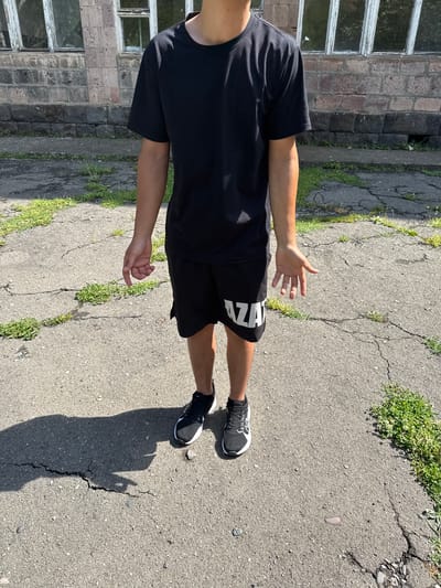 A person stands in front of a brick building in Tumanyan, Armenia. The building has windows with white frames and appears to be old. The ground in front of the building is paved with asphalt and has cracks with grass growing through them. The person is wearing a black t-shirt, black shorts with white lettering, and black and white sneakers. Their shadow is cast on the ground to their left. The person's right hand is outstretched.