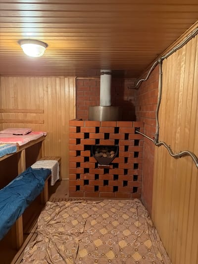 The image shows the interior of a Russian banya (sauna) in the city of Kamenka. The walls and ceiling are paneled with light-colored wood. A brick stove with a metal chimney is centrally located, with stones visible inside its lower section. To the left, wooden benches are covered with towels and a blue tarp. In the foreground, a patterned blanket covers a wooden floor. A ceiling light fixture is visible. Metal pipes run along the brick wall.