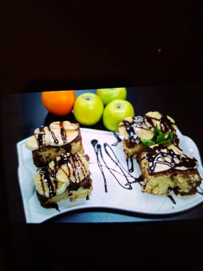 The image displays a plate of four dessert pieces, each topped with cream and drizzled with chocolate sauce. Behind the plate are three green apples and one orange. The desserts appear to be some type of cake or pastry. The background is dark and out of focus. The scene is set indoors on a dark surface. There is no indication of the location in Medvezhyi Ozyora, Russia, or any people.