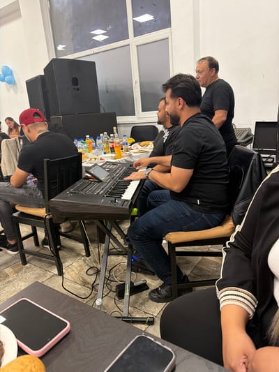 The image captures an indoor event, possibly a musical performance or gathering, in the city of Dalgopol, Bulgaria. In the foreground, a man wearing a red baseball cap and a black t-shirt is seated and looking towards a keyboard, his hands positioned as if playing. Beside him, another man in a black t-shirt is also seated and looking towards the keyboard. Behind them, two men stand. One, also in a black t-shirt, is looking away from the camera. The other, an older man in a black t-shirt, is looking to his right. A large, black electronic keyboard with numerous buttons and keys is set up on a stand in the center of the frame. Bottles of drinks and food items are visible on a table in the background, and large black speaker boxes are stacked to the left of the scene. Blue balloons are attached to a wall on the far left. The lighting suggests an indoor space with large windows reflecting overhead lights.