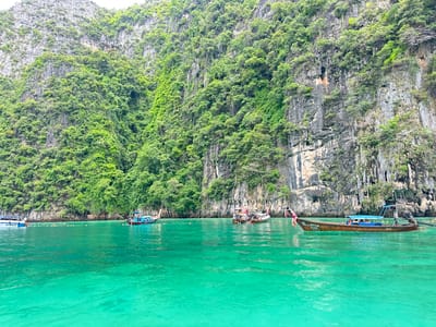 This image depicts a scene in the city of Haad Yao, Thailand, characterized by towering limestone cliffs covered in lush green vegetation, dropping sharply into crystal-clear turquoise water. Several boats are visible in the water. In the foreground, a longtail boat with a blue canopy is prominently featured, carrying passengers. To the left, another longtail boat is anchored. Further in the distance, a larger white boat with "BLUE MARINE 255" visible on its side is present. A rope with buoys stretches across the water, separating areas. A few individuals can be seen in the water between the boats, some appearing to be swimming or wading. The water exhibits transparency, allowing glimpses of the seabed in some areas. The overall environment suggests a tropical coastal location, likely a popular tourist destination.