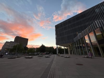 The image depicts an urban scene in Karlsruhe, Germany, specifically in front of the KIT (Karlsruhe Institute of Technology) South Campus, often referred to as the "Kaiserstraße" campus. The photograph was taken at dusk, with a vibrant sunset casting warm hues of pink and orange across the sky, contrasting with the pale blue.
In the foreground, a wide, paved plaza with a grid-like pattern of light grey tiles dominates the scene. Several modern, angular concrete benches are strategically placed throughout the plaza. A drainage grate runs horizontally across the middle of the plaza, leading the eye towards the buildings.
To the left, a tall, rectangular building with many windows and a dark facade stands at an angle. Its upper portion features a unique, propeller-like structure. To the right, a larger, contemporary building with a dark, ribbed facade and extensive glass windows is prominently featured. The glass reflects the colorful sunset and the surrounding environment.
Trees with green foliage are visible in the middle ground, lining the street behind the plaza. In the background, more buildings and streetlights can be seen. A person on a bicycle is captured in motion on the left side of the plaza, adding a sense of activity to the otherwise still scene. Another bicycle is parked near the modern building on the right. The overall atmosphere is serene, with the dramatic sunset illuminating the cityscape.