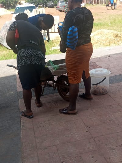 Three individuals are observed outdoors on a reddish-brown patterned paved surface in Ajalli, Nigeria. A dark, single-wheeled wheelbarrow is positioned centrally. To the left, a female wearing a black and white patterned top and dark skirt, with a red bag slung over her left shoulder, is bent over the wheelbarrow. Adjacent to her, a male in a blue shirt is also bent over the wheelbarrow, seemingly interacting with its contents and holding a clear plastic item. To the right, a second female in a black patterned top and orange skirt stands upright, facing away from the viewer; a dark bag and a blue and black striped object are slung over her shoulder. All individuals wear sandals, except for the male who wears closed-toe shoes. A clear plastic bucket containing several brown, round objects rests on the pavement to the right of the wheelbarrow. The background features an unpaved dirt area with grass, several parked vehicles including a white SUV, and piles of earth and building blocks.