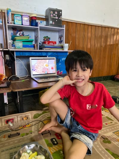 The scene is an indoor room in Đak Đoa, Vietnam. A young East Asian boy, approximately 5-8 years old, is seated on a multi-colored playmat featuring roads and buildings. He is barefoot, wearing a red short-sleeved t-shirt with a white "Zalo" logo and faded blue denim shorts. He has short black hair, a silver-colored chain necklace, and a silver-colored bracelet on his right wrist. The boy is smiling broadly, exposing his teeth, and has his right hand resting on his chin.
In the bottom left foreground, a silver-colored fluted metal bowl contains several slices of light-colored fruit, likely guava.
Behind the boy is a wooden desk with blue decorative elements and white shelves. On the desk, a silver-colored Apple MacBook laptop is open and displaying a colorful, cartoon-style video game or animation. To the left of the laptop, a black smartphone rests on the desk, and a black microphone on an articulating arm is clamped to the desk's edge.
The white shelves above the desk contain various items. The top shelf displays a blue and a purple handheld electronic device, a red and black container, and a clear bottle. The bottom shelf holds several books, a green pencil case, a blue container labeled "Ovaltine," a red box of "Toffee's," additional papers, and a white mug with "TẾT 2020" inscribed. Above the shelves, a grey metal safe is affixed to the wall, adorned with multiple magnets and stickers. The wall behind the desk is partially covered by light brown wooden paneling at the bottom, transitioning to a light cream-colored painted surface with faint pencil or crayon scribbles visible on the upper section. Beyond the playmat, a dark tiled floor is visible.