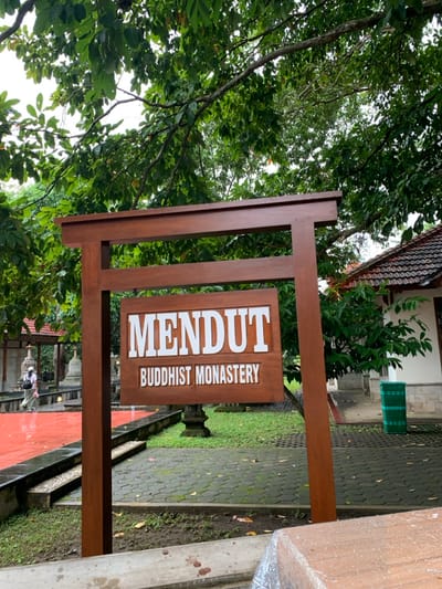 The image shows a wooden signpost with the words "MENDUT BUDDHIST MONASTERY" on it. The sign is located in the city of Mungkid, Indonesia, at the coordinates -7.605 latitude and 110.23 longitude. The sign is standing on a paved walkway in front of a lush green lawn. The sign is partially obscured by a tree. There is a person in the background, walking towards the sign. The sign is in a traditional Indonesian architectural style, with a brown wooden frame and a white inscription. The background is a green lawn with a red brick pathway. There is a building in the background to the right. It is a white building with a red tiled roof. There is a green trash can next to the building. The person is walking on the red brick pathway, heading towards the sign. The environment is peaceful and serene, with lush green vegetation and a clean, well-maintained paved walkway.