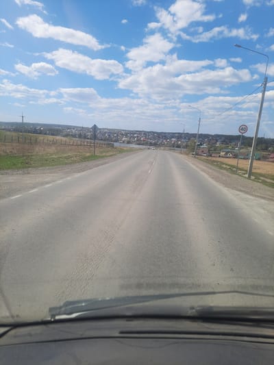 A long, straight asphalt road stretches into the distance, seemingly leading into the heart of a town. The road is bordered on one side by a field with utility poles and fences and on the other by a settlement. The settlement appears to be the town of Научный Городок, Russia. Streetlights line the road, and a speed limit sign with the number "40" is visible. The sky above is a bright blue, filled with fluffy white clouds. The image is taken from within a vehicle, as part of the windshield and wiper are visible in the foreground. The weather appears to be clear and sunny.