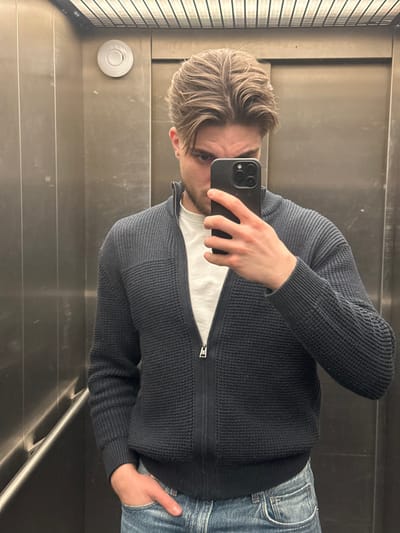 The scene is an elevator located in Volketswil, Switzerland. A male individual, appearing to be in his 20s to 30s, is captured in a mirror selfie. He has light brown, styled hair and displays a neutral facial expression with his mouth and nose obscured by a black smartphone held in his left hand. He is dressed in a white crew-neck t-shirt, an unzipped dark blue/grey ribbed knit cardigan, and blue denim jeans. His right hand is placed inside his right jeans pocket. The elevator interior consists of reflective metal walls with visible vertical panel seams and surface imperfections. A circular white object, identified as a smoke detector or emergency light, is mounted on the upper left wall. The ceiling features a metallic grid pattern. A horizontal handrail or reflective strip is present on the lower left wall.
