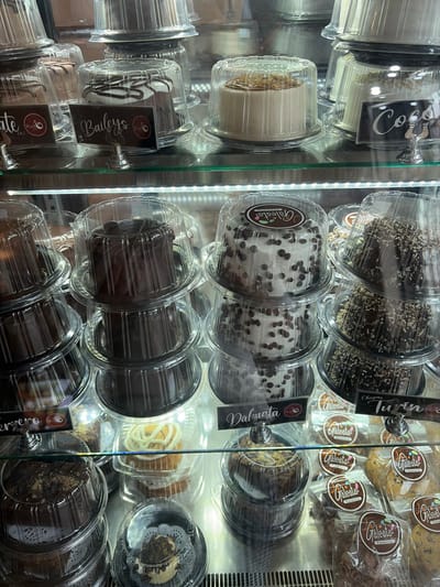 A refrigerated display case showcases various cakes. Top rows feature "Coco" and "Turin" cakes. The second row contains "Valnata" cakes with white frosting and chocolate chips. Below, chocolate cakes sit in rows. Cookies and pastries are also visible. Each cake is individually packaged in clear plastic containers with labels.