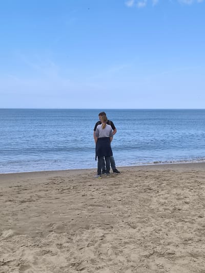 Two people, one man and one woman, are standing on a sandy beach, facing the water. The man is wearing a dark-colored shirt, and the woman is wearing a light-colored shirt with a dark jacket around her waist. They appear to be conversing. The beach is sandy, and the sea is calm with small waves. In the distance, there are multiple structures that appear to be wind turbines. The sky is clear and blue, suggesting a bright day. The image was taken on a beach in Bergen, Netherlands, as indicated by the presence of wind turbines.