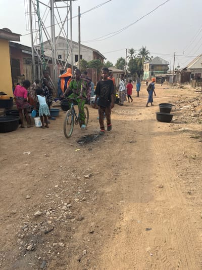 The image displays an outdoor scene on an unpaved, dusty street in Makera, Nigeria, under a clear, light blue sky. The ground is characterized by compacted dirt, loose gravel, and scattered debris, with visible tire tracks and dark, burnt patches.

On the left side of the street, adjacent to a utility pole, stands a tall metal lattice structure consistent with a water tower or similar utility installation. Below this structure, a group of several individuals, predominantly women and children, are gathered. They are interacting with multiple large, black plastic tubs and smaller colorful buckets, indicative of water collection or distribution activities. Notable figures include a woman in a pink top and a child in a light blue dress within this group.

In the center foreground, an individual wearing a vibrant green patterned outfit is riding a light blue bicycle, moving from the left side of the frame towards the right. Directly beside and slightly ahead of the bicyclist, a young male in a dark t-shirt and patterned trousers walks in the same direction.

Further down the street on the right, two additional large, black plastic tubs are positioned on the ground. The background is composed of various residential structures lining both sides of the street, some appearing unfinished or weathered. Scattered individuals are visible walking or standing in the mid-to-far background. Overhead utility lines and palm trees are also present within the scene.