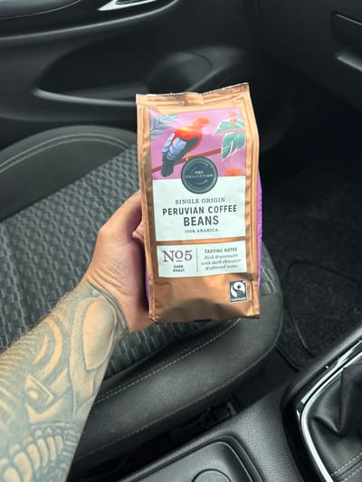 This image shows a person's hand holding a bag of M&S Collection Single Origin Peruvian Coffee Beans. The bag has a purple and gold design featuring an illustration of a bird and tropical leaves. Text on the bag indicates it is 100% Arabica, dark roast, with tasting notes of rich and aromatic, dark chocolate, and almond. The Fairtrade logo is also visible. The scene is set inside a car, with dark upholstery visible on the seats and part of the gear stick in the background. The person's tattooed arm is also visible, extending into the frame. The lighting suggests it is daytime. The context provided indicates the location is Newcastle-under-Lyme, United Kingdom.