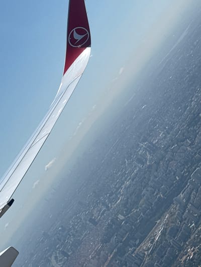 The image shows a view from an airplane window, looking down at a dense urban area. A portion of the airplane's wing, which is white and red and features a logo, is visible in the foreground. The city below is characterized by numerous buildings and a network of streets. The atmosphere is hazy, obscuring some of the finer details of the cityscape. The angle of the photograph suggests the airplane is in flight, likely during descent or ascent, given the proximity to the urban landscape. Based on the provided context, this scene is likely depicting the city of Avcılar, Turkey.