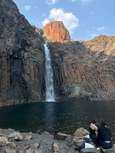 The media file depicts a scenic view of a waterfall cascading down a rocky cliff face. Several people are visible in the scene, either near the top of the cliff, appearing to be preparing for an activity, or seated on rocks by the water's edge. One individual is suspended mid-air, suggesting a bungee jump or similar recreational activity is underway. The environment is characterized by rugged, dark, and reddish-brown rock formations. A body of dark, rippling water is situated at the base of the waterfall, with rocky terrain surrounding it. The sky above is partly cloudy with a blue hue. The lighting suggests it is daytime. The overall atmosphere seems to be one of adventure and natural beauty.
