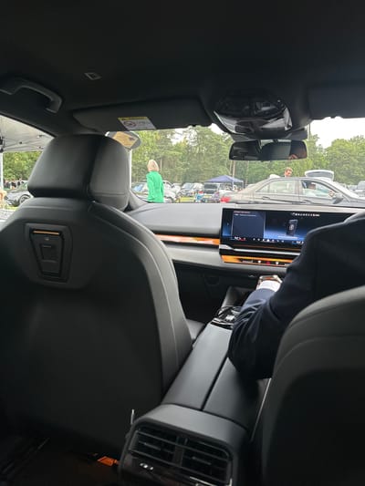 A photograph taken from inside a car shows a person's hands and arm resting on a center console. The car's dashboard display is visible, showing the text "CONNECT NEW DEVICE" and a Bluetooth symbol. Outside the car, a woman in a bright green jacket is standing near a parked car, with other vehicles and trees in the background. The scene appears to be at an outdoor event or gathering, possibly a car show, with tents and parked cars visible. The location is specified as Ascot, United Kingdom.