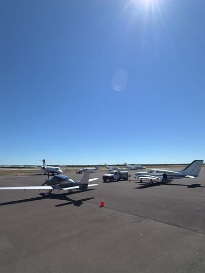 This is a wide-angle, eye-level, outdoor, daylight, full shot of an airport tarmac in the Town of Southampton, United States of America. The sky is a clear, bright blue, with the sun visible in the upper right corner, creating a lens flare. The tarmac is asphalt. In the foreground, a small, single-engine airplane with a silver fuselage and white wings is parked, facing right. Its registration number appears to be N132JW. To the right of this airplane, a larger, twin-engine propeller aircraft with a white fuselage and dark green trim is parked, facing right. Its registration number is 42AA. Behind these two planes, further down the tarmac, are several other aircraft, including a white jet airplane with a dark stripe on its tail. A small white truck with a fuel tanker trailer is parked to the left of the twin-engine propeller aircraft. Several orange traffic cones are placed on the tarmac around the parked vehicles and aircraft. The background consists of a flat, treeless landscape. There are no people visible in the image.