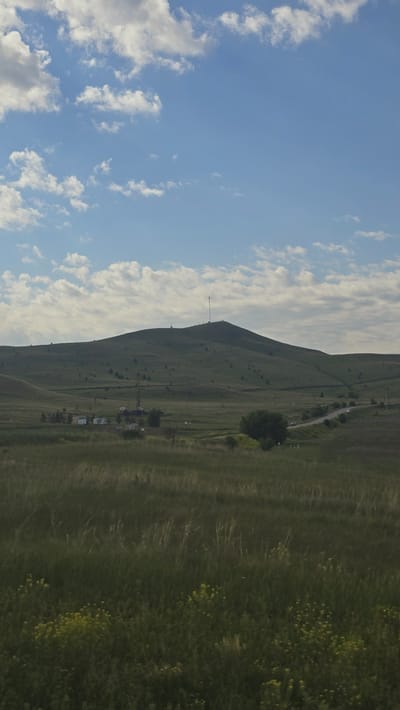 The image shows a vast, rolling landscape under a partially cloudy sky. In the foreground, tall, green grass dominates, with scattered patches of small yellow wildflowers. A few trees are visible in the middle ground, along with what appears to be a drilling rig and several small buildings, suggesting some industrial activity. Further in the distance, a large, rounded hill rises, its slopes covered in a mix of grass and sparse trees. A prominent communications tower stands atop the hill, along with smaller structures. A winding road snakes across the middle ground, with a few cars visible on it. The sky is a clear blue with scattered white cumulus clouds. The overall scene depicts a rural or semi-rural environment with a mix of natural landscape and man-made structures.