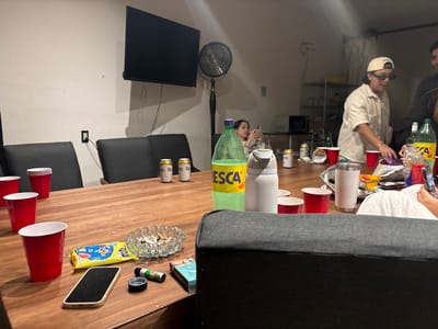 A cluttered wooden table sits in a room with a TV, fan, and shelving in the background. Red plastic cups, beer cans, a green soda bottle, and a white water bottle are scattered across the table. An ashtray full of cigarette butts, a phone, and cigarette packs are also present. Two people stand around the table, while another person sits in the background looking at a phone. The room appears to be in a state of disarray.